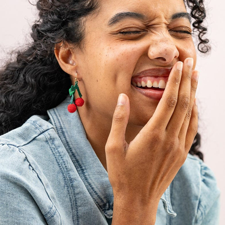 Woman wearing colorful earrings and a bracelet, laughing with her hand on her face against a light pink background