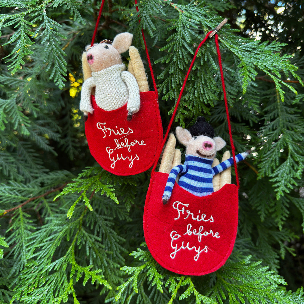 Two knitted pig ornaments in red sacks with text, hanging on a tree.