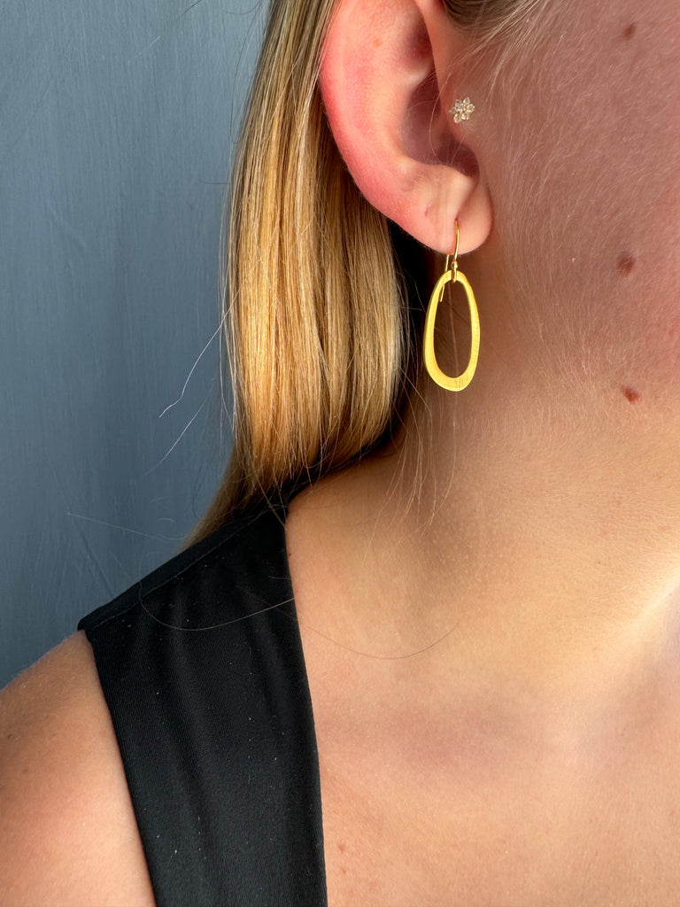 Gold earring being worn by a person with blonde hair and a black top on a neutral background.
